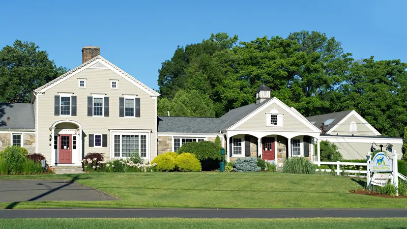 Cream colonial building with red doors, a covered porch and a sign, set behind a green lawn on a sunny day