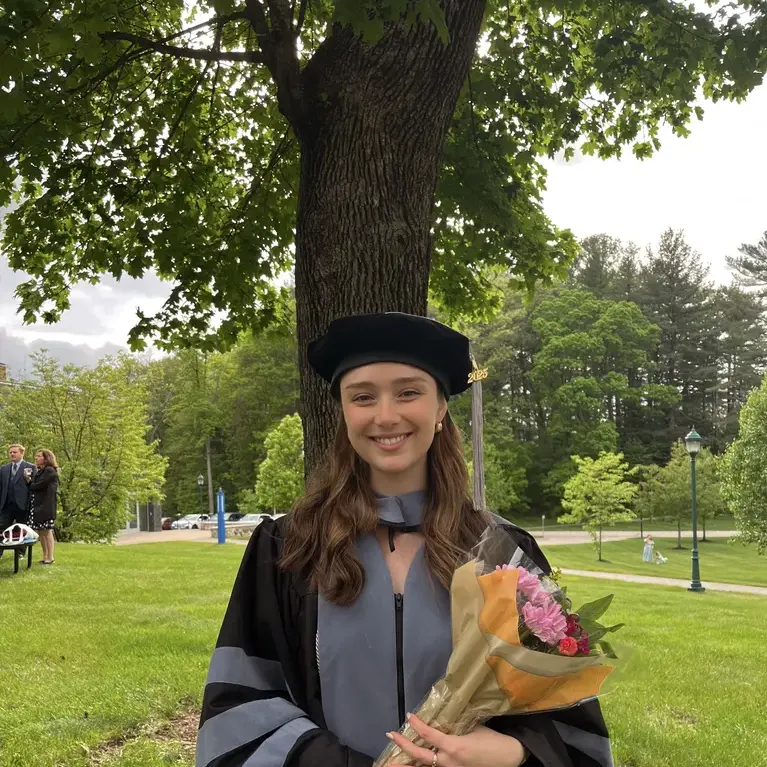 Tara Francis, DVM, MS: Smiling woman in a doctoral graduation gown and cap holding a bouquet under a large tree